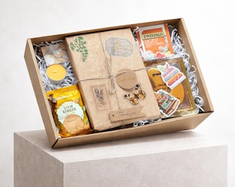 May include: A gift box filled with various items, including a wrapped package tied with twine, a tea box, a jar of honey, a candle, and a bag of stem ginger cookies. The box is lined with white paper and decorated with stickers and a butterfly.