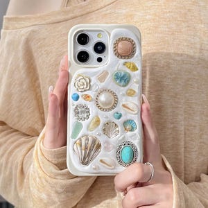 May include: White iPhone case decorated with various seashells, pearls, and turquoise stones. The case has a raised design and a camera lens cutout.