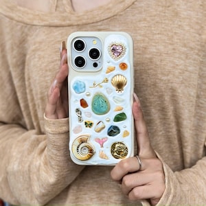 Könnte beinhalten: Cremefarbene Handyhülle mit einer Vielzahl dekorativer Elemente. Die Hülle verfügt über einen herzförmigen Schmuckstein, Muscheln, Edelsteine und andere Verzierungen. Die Hülle hat ein Mehrkamerasystem.