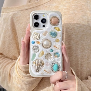 Può includere: Custodia per telefono bianca decorata con varie conchiglie, perle e pietre preziose. La custodia presenta una varietà di colori, tra cui bianco, oro, blu e rosa. Il design include una grande perla, una pietra turchese e un piccolo fiore.