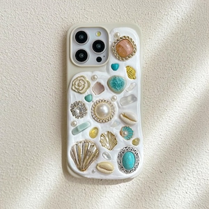 Può includere: Una custodia per iPhone bianca decorata con vari abbellimenti, tra cui conchiglie, perle, strass e pietre turchesi.
