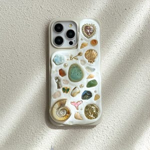 May include: A beige iPhone case adorned with various decorative elements. The case features a heart-shaped jewel, seashells, gemstones, and a gold coin. The design includes a mix of textures and colors, creating a unique and artistic aesthetic.