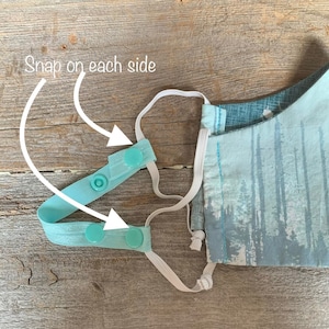 May include: A light blue face mask with a white elastic band and two turquoise snap buttons on each side. The text "Snap on each side" is written on the image.