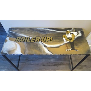 May include: A white and brown resin table top with a black metal frame. The table top has a marbled design and features a gold graphic of a cartoon figure with the text "BOILER UP!"