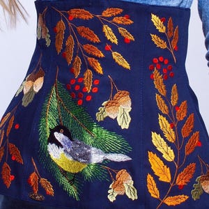 以下が含まれることがあります： 青い布地のコルセットに、刺繍された秋の葉、ベリー、小さな鳥が描かれています。鳥は黄色、灰色、黒で、長い尾をしています。