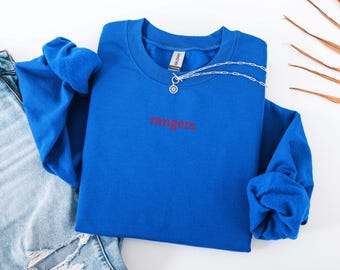 Puede incluir: Una sudadera azul real con la palabra "rangers" bordada en rojo. Un collar plateado con un colgante está drapeado sobre la sudadera. La sudadera está doblada sobre un par de vaqueros azules.