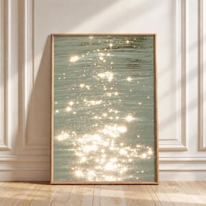Può includere: Una stampa artistica da parete rettangolare con orientamento verticale. L'immagine mostra uno specchio d'acqua color verde salvia con luminosi riflessi bianchi a forma di stella sparsi sulla superficie increspata, incorniciata in legno chiaro.