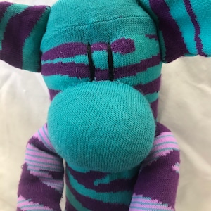 May include: A handmade sock monkey doll in shades of teal, purple, and lavender. The monkey has a teal head and snout, with purple and lavender striped arms, legs, and body. The ears are teal with purple stripes. The doll is made from socks.