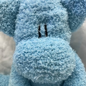 Puede incluir: Primer plano de un elefante de peluche azul claro con ojos cosidos negros. El elefante tiene orejas grandes y una superficie texturizada y difusa con detalles de purpurina plateada. El juguete es suave y tierno.