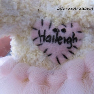 May include: A plush, cream-colored stuffed animal with a pink heart-shaped patch. The patch is embroidered with the name "Haileigh" in black. A pink tulle skirt is visible at the bottom of the image.