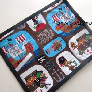 Puede incluir: Un artículo de tela cuadrada marrón con un borde negro. La tela presenta un patrón repetido de barcos piratas, cañones, cofres del tesoro y personajes piratas en varias formas. El fondo es marrón con acentos blancos y azules.