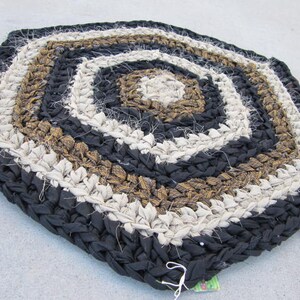 May include: Handmade, hexagonal rag rug in a spiral pattern. The rug features concentric bands of black, tan, off-white, and light brown fabric strips. The rug is made of braided fabric strips and is suitable for a variety of home decor styles.