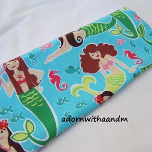 Puede incluir: Una funda rectangular de tela turquesa con un estampado de sirenas. El diseño incluye sirenas con cabello castaño, colas verdes y tops de conchas rosas, junto con caballitos de mar, peces y otros elementos acuáticos. El texto "adornwithaandm" es visible.