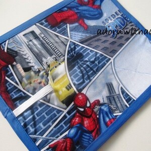 Puede incluir: Un agarrador de ollas cuadrado y acolchado con un diseño de cómic que presenta a Spider-Man en poses de acción. El fondo incluye paisajes urbanos y el texto "SPIDER". El agarrador tiene un borde azul.