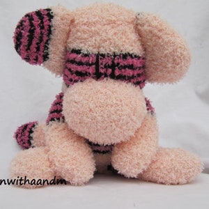 May include: A plush pink dog toy with black and pink striped accents on the ears, paws, and around the neck. The toy is sitting down with its legs extended. The text "adornwithaandm" is visible in the bottom left corner.