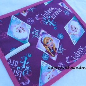 以下が含まれることがあります： ピンクの縁取りが付いた正方形の紫色の布製品で、アニメのキャラクターの画像と「Sisters Forever」の文字が特徴です。デザインには雪の結晶とひし形の肖像画が含まれています。白いハンドルが付いています。