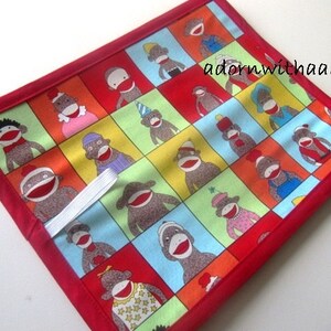 May include: A square, quilted potholder with a patchwork design of sock monkey faces in different outfits. The squares are set against a red border. A white elastic loop is attached for hanging. The text "adornwithaandm" is visible.