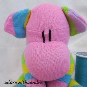 May include: A handmade pink sock animal with rainbow-striped ears and legs. The animal has black stitched eyes and a stitched mouth. A spool of blue thread sits next to the animal. The text "adornwithaandm" is at the bottom.