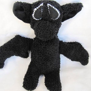 May include: A black plush bat toy with large ears and outstretched wings. The bat features white stitched eyes and a soft, textured body. This cuddly toy is ideal for Halloween or as a unique gift.