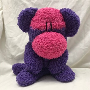 May include: A plush dog toy with a purple body, ears, and paws, and a pink face and nose. The toy has a soft, textured appearance and is sitting upright. Black stitching details the eyes and mouth.