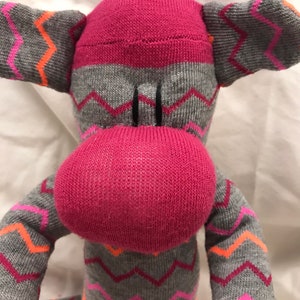 May include: A handmade sock monkey doll. The monkey has a pink head and nose, with gray and pink zigzag patterned arms, legs, and ears. The monkey's eyes are black. The doll is made from socks.