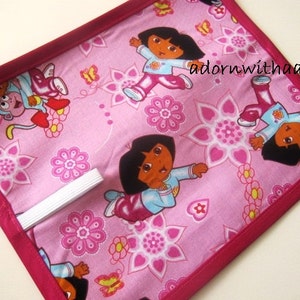 May include: Pink fabric item with a repeating pattern of Dora the Explorer characters and floral designs. The fabric is bordered with a darker pink trim. A white elastic band is visible. The text "adornwithaandm" is in the upper right corner.