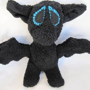 May include: A black plush bat toy with large ears and outstretched wings. The bat's eyes are outlined in blue stitching. The toy is made of a soft, textured material.