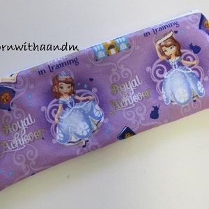 May include: A lavender pencil case with a zip. The case features a repeating pattern of a princess in a blue dress, the words "Royal Achiever" and "in training". There are also images of castles and books on the case.
