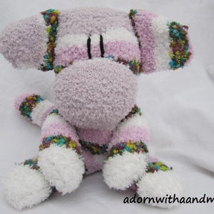 May include: A handmade plush sock monkey in shades of lavender, white, and pink. The monkey has striped arms, legs, and body, with white paws and ears. The eyes are black, and the nose is a lighter shade of lavender. The text "adornwithaandm" is visible.