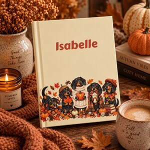 May include: A cream-coloured hardcover book with the name Isabelle printed in red. The cover features an illustration of five dachshunds surrounded by autumn leaves, pumpkins, and acorns. The dogs are wearing sweaters and scarves, with one holding a pumpkin.