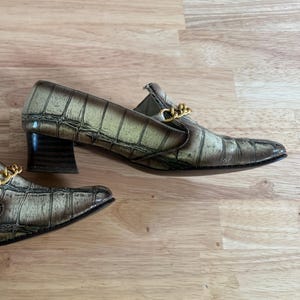 May include: A pair of brown and tan faux-crocodile leather loafers with a gold chain detail across the vamp. The shoes have a block heel and a pointed toe. The shoes are displayed on a wooden surface.