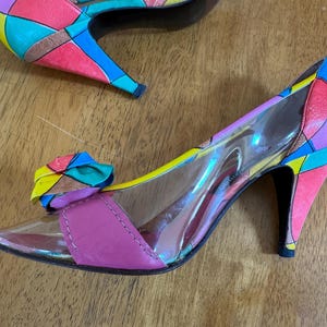 Op de afbeelding: Een paar kleurrijke pumps met een doorzichtige vamp en een strikdetail. De hakken en het bovenste deel van de schoenen zijn versierd met een geometrisch patroon in verschillende kleuren, waaronder roze, geel en blauw.
