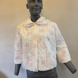 May include: A white quilted jacket featuring a delicate floral pattern in soft pink and light blue. The jacket has three-quarter length sleeves, a rounded collar trimmed with white lace, and a front button closure with two visible buttons.