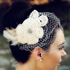 May include: Ivory crochet flower and veil headpiece. The headpiece features a large, detailed crochet flower with a rhinestone center, accented by feathers. A delicate net veil drapes over the face, creating an elegant bridal accessory.
