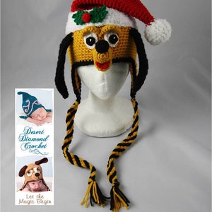 Op de afbeelding: Een gebreide muts met een geel honden gezicht, zwarte oren en een rode kerstmanmuts erop. De muts heeft een lange zwart-gele gestreepte stropdas. De muts zit op een wit mannequin hoofd. De afbeelding bevat ook een logo voor "Desert Diamond Crochet" met de tekst "Let the Magic Begin".