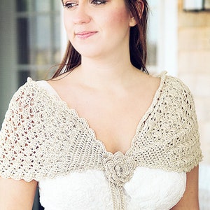 Pode incluir: Uma mulher vestindo um vestido branco com um xale de crochê. O xale é bege e tem um desenho floral no centro.
