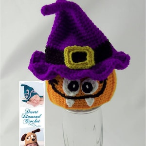 May include: A crocheted pumpkin hat with a purple witch hat on top. The pumpkin hat has a smiling face with white teeth and black eyes. The witch hat has a black band with a gold buckle. The hat is sitting on a clear glass jar. The text "Desert Diamond Crochet" and "Let the Magic Begin" is visible on the image.