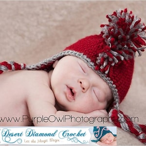 May include: A newborn baby wearing a red and gray crocheted hat with a pom-pom on top. The hat has a long tail that is braided with red and gray yarn. The baby is sleeping peacefully on a beige surface. "Desert Diamond Crochet Let the Magic Begin..." is written on a banner below the baby.
