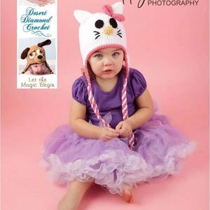 May include: A young child wearing a white crocheted cat hat with pink ears and a pink bow. The hat has black eyes and a yellow nose. The child is wearing a purple shirt and a lavender tulle skirt. The background is a light pink.