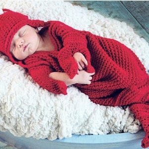 May include: A newborn baby wearing a red crocheted lobster costume, sleeping in a blue wooden boat on a white fluffy blanket.