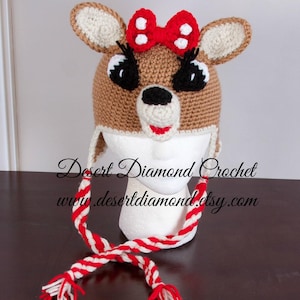 May include: Crocheted deer hat with a red bow and white and red striped scarf. The hat is brown with black eyes and a white nose. The text "Desert Diamond Crochet www.desertdiamond.etsy.com" is visible on the hat.