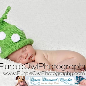 May include: A newborn baby wearing a green crocheted hat with white polka dots, sleeping on a white fluffy blanket. The hat has a long pointed top.