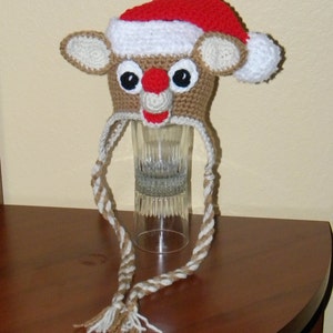 Op de afbeelding: Een gehaakte rendiermuts met een rode en witte kerstmanmuts erop. De muts heeft een bruin gezicht met zwarte ogen en een rode neus. De muts heeft een lang bruin en wit gehaakt koord.