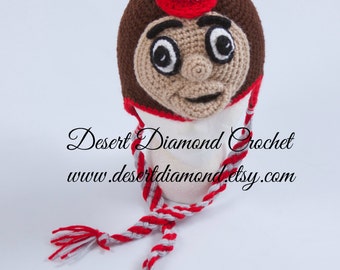 May include: Crocheted hat featuring a cartoon face with a red and white striped scarf. The hat has a gray and white baseball cap on top with a red "O" on the front. The text "Desert Diamond Crochet www.desertdiamond.etsy.com" is visible below the hat.