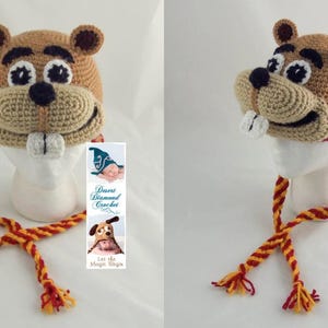 May include: A crocheted brown and tan hat shaped like a cartoon beaver with a toothy grin. The hat has long red and yellow ties. The hat is on a white mannequin head. The hat is accompanied by a small card with the text "Desert Diamond Crochet" and "Let the Magic Begin".