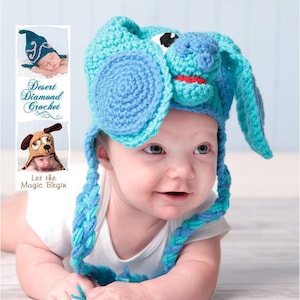 Op de afbeelding: Een baby draagt ​​een blauwe en turquoise gehaakte muts met een hondenkop. De muts heeft lange, gevlochten banden. De baby ligt op zijn buik en kijkt naar rechts.