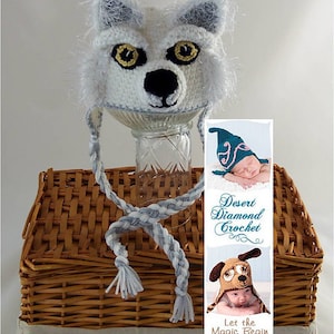 Pode incluir: Chapéu de lobo branco de crochê com laços trançados cinza e branco. O chapéu é exibido em uma cesta de vime com um cartão que diz "Desert Diamond Crochet" e "Let the Magic Begin".
