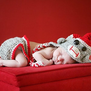 Può includere: Un bambino indossa un cappello da scimmia all'uncinetto grigio e rosso e un pannolino abbinato e dorme su un cuscino rosso.