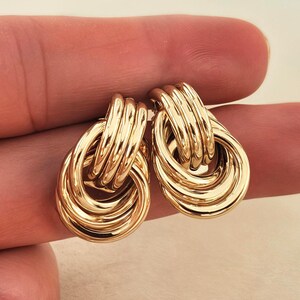May include: A pair of gold-colored earrings. Each earring features a looped design with multiple stacked, ridged elements. The earrings have a polished, reflective surface, and are shown against a neutral background.
