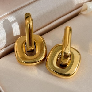 Peut inclure: Une paire de boucles d'oreilles dorées. Chaque boucle d'oreille présente une base carrée aux bords arrondis et une boucle supérieure rectangulaire. Les boucles d'oreilles sont présentées sur une surface satinée de couleur claire, mettant en valeur leur finition polie.